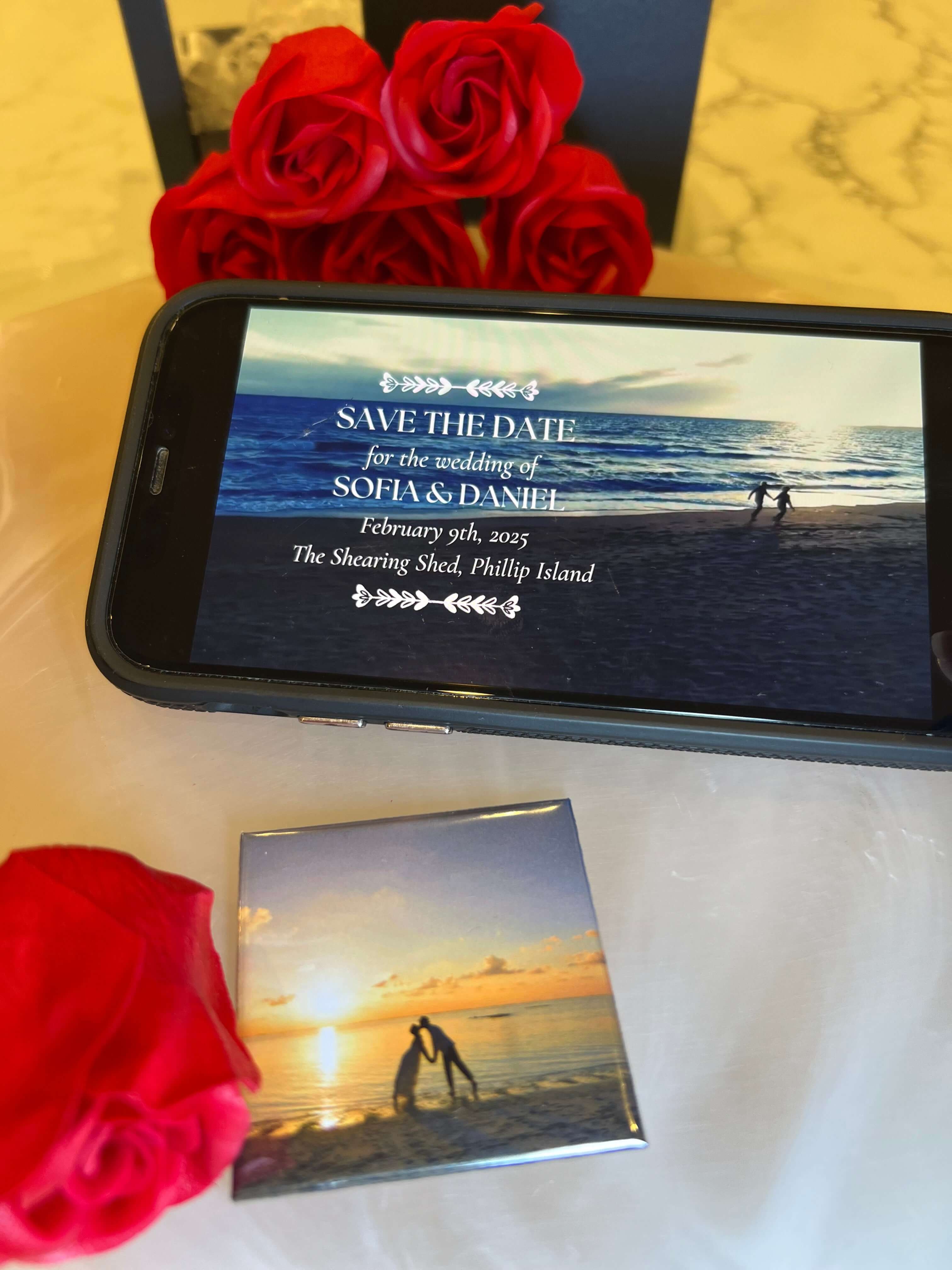 Phone displaying a wedding invitation with red roses and a mirror reflecting the scene on a marble surface. Custom photo magnets. Tap & Play Magnets.