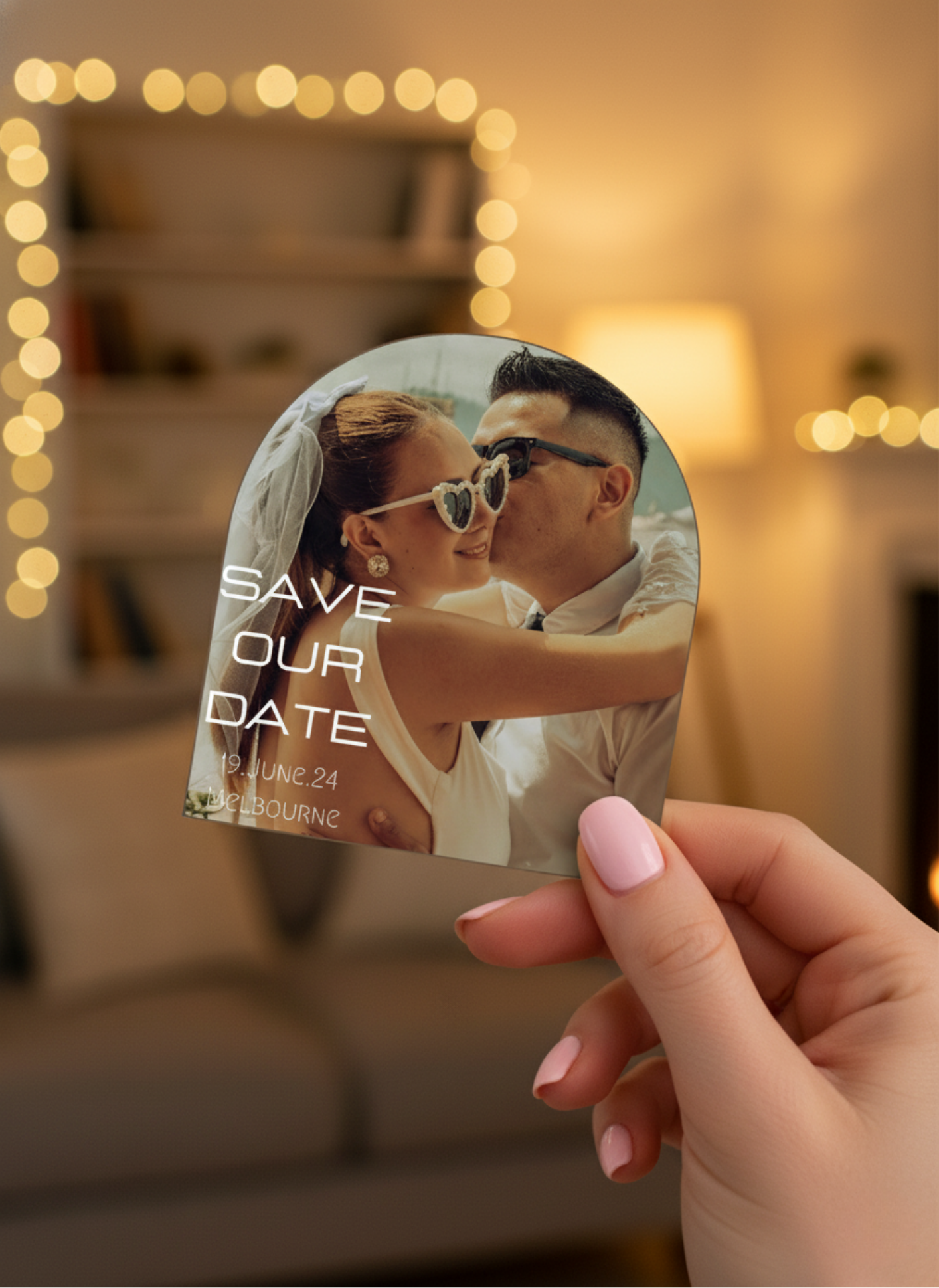 Hand holding a 'Save the Date' card with a couple kissing, blurred lights in the background