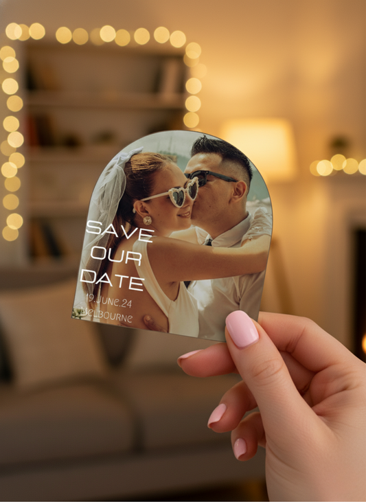 Hand holding a 'Save the Date' card with a couple kissing, blurred lights in the background